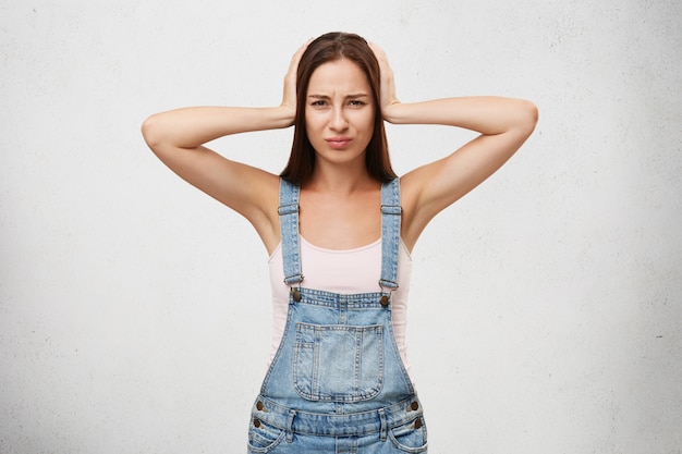 Beautiful female in denim overalls suffering from loud noise and headache, covering head and ears with hands, looking upset isolated. Human emotions, feelings and reaction