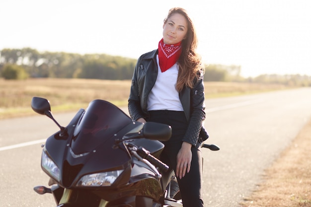 Beautiful female driver sits on black fast motorbike, dressed leather jacket, travels around country by motorbike, stops on side