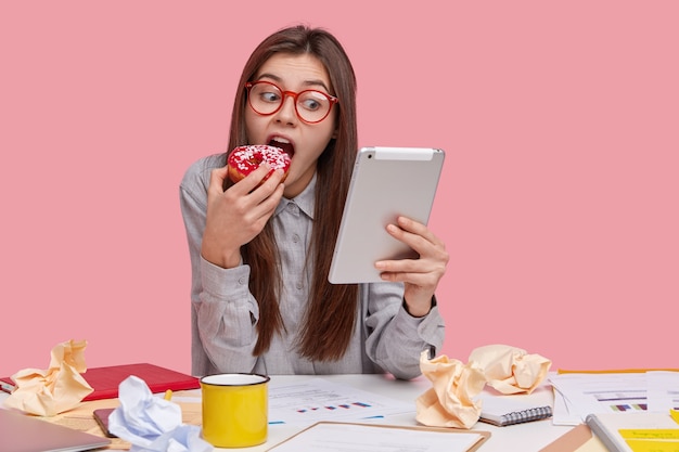 Free photo beautiful female freelancer has coffee break after studying documentation, holds tablet, watches movie online, bites tasty homemade doughnut