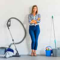 Free photo beautiful female housemaid with folded hand standing near cleaning equipments