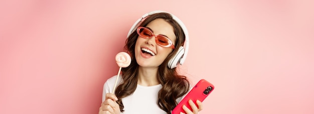 Free photo beautiful female model listening music in headphones holding lolipop and mobile phone posing in sung