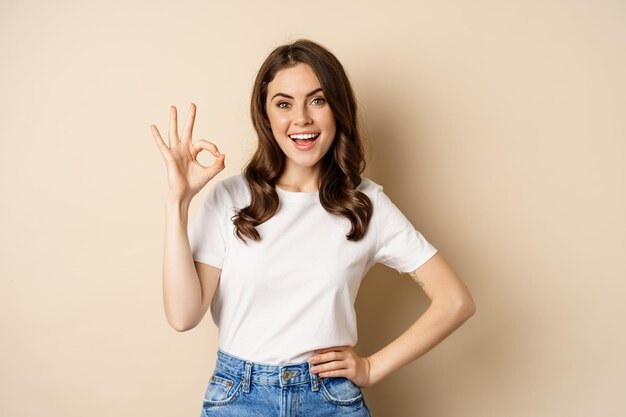 Beautiful female model smiling pleased, showing okay, zero sign, complimenting, praising you, standing over beige background