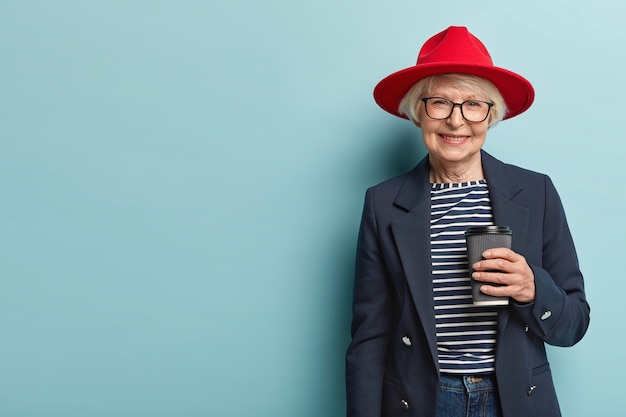 Free Photo beautiful female pensioner has meeting with colleague, discusses something during coffee break, holds paper cup of cappuccino, wears stylish outfit, isolated on blue wall, copy space on left