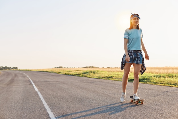 Beautiful female wearing t shirt, short and hair band skateboarding in the street, looking in distance, spending time alone with pleasure, active and healthy lifestyle.