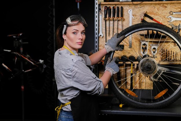 Beautiful female in working clothes, apron and goggles, repairs a bicycle wheel in a workshop.