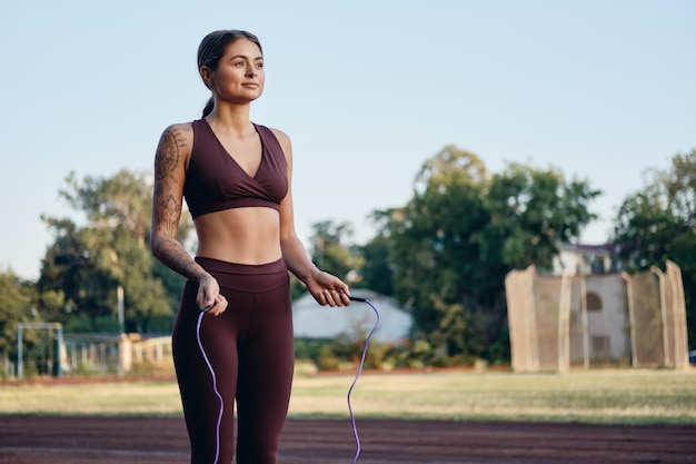 Beautiful fitness girl in stylish sportswear with skipping rope dreamily tranining on city stadium