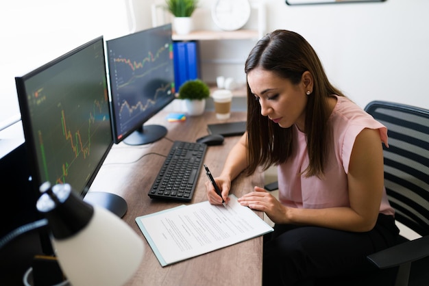 Beautiful freelancer and stock broker signing a business deal at her work desk. Successful businesswoman reading a work contract