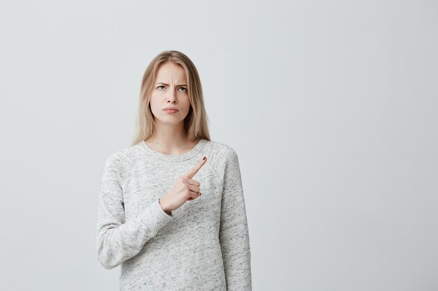 Beautiful furious woman with blonde dyed hair looking sidewards with serious and angry expression pointing index finger, showing copy space