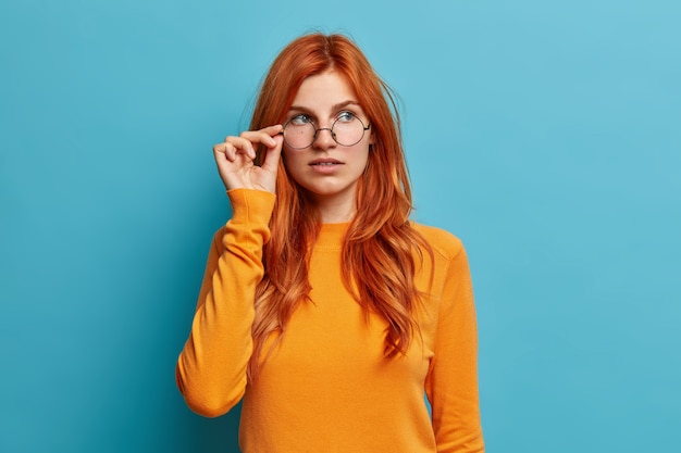 Free photo beautiful ginger female student thinks about future project keeps hand on spectacles concentrated pensively aside dressed in casual orange jumper.