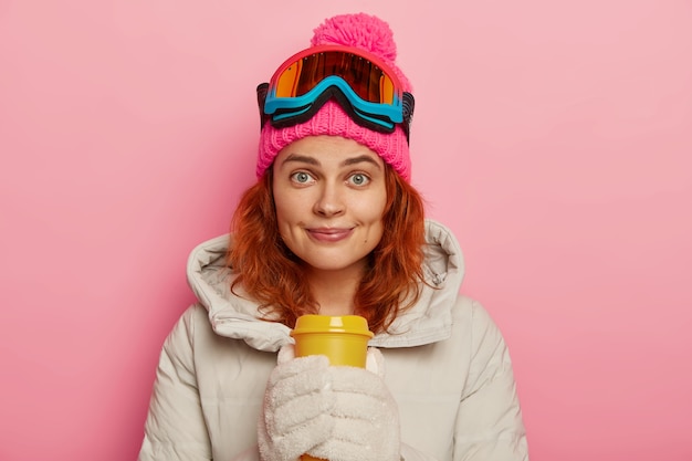 Free photo beautiful ginger female teenager enjoys extreme sport, drinks coffee after snowboarding, looks with pleasure at camera, wears ski goggles