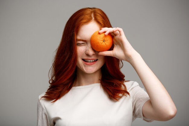 beautiful ginger girl holding orange over gray wall.