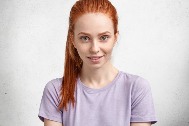 Free photo beautiful ginger woman in casual purple t shirt, looks with pleased delighted expression at camera, poses against white concrete wall.