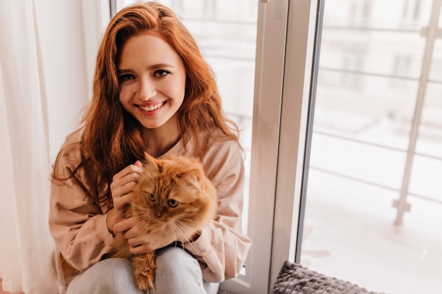 Beautiful ginger woman posing with smile beside window. Indoor shot of attractive girl holding red fluffy cat.