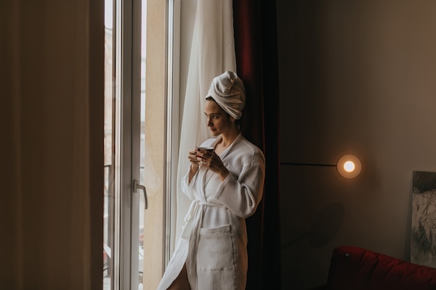 Free photo beautiful girl in bathrobe and towel on her head looks thoughtfully out window with cup of tea in her hands.