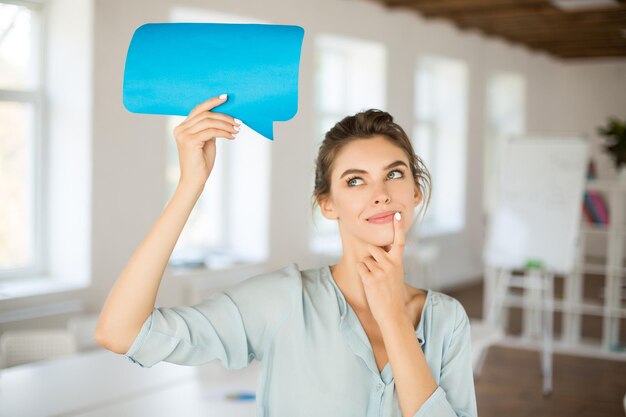 Beautiful girl in blouse dreamily looking aside holding blue paper message icon in hand near head while spending time in office