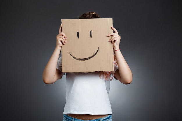 Beautiful girl hiding behind carton with smiley over grey wall.