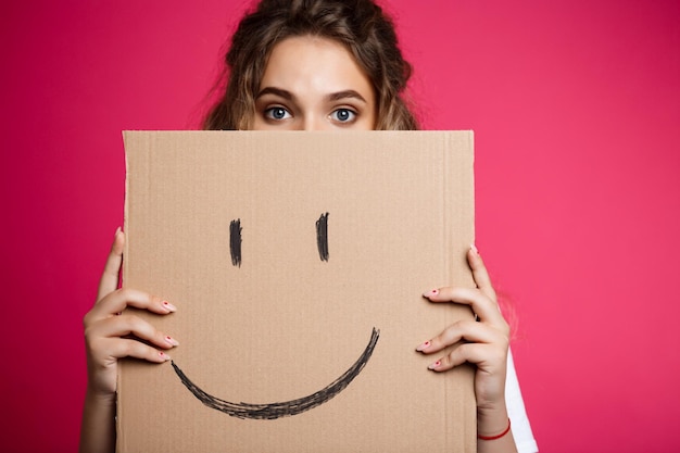 Beautiful girl hiding behind carton with smiley over pink background