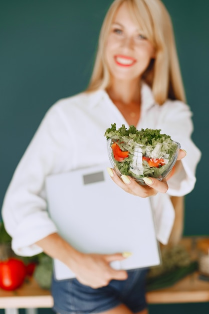 Beautiful girl make a salad. Sporty blonde in a kitchen. Woman holding weights in her hands.