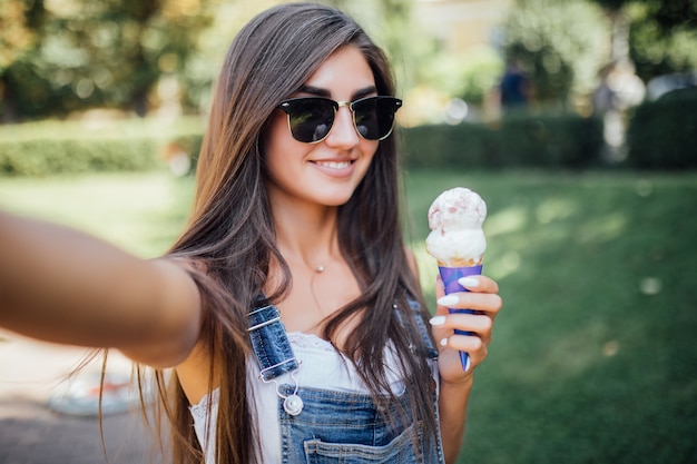 Beautiful girl makes selfie smiles with white teeth and holds the ice cream wearing sunglasses