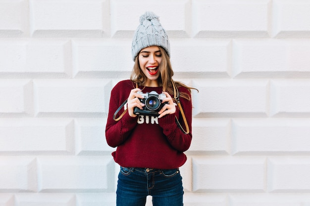 Free photo beautiful girl in marsala sweater on grey wall . she wears knitted hat, is astonished looking at camera in hands.