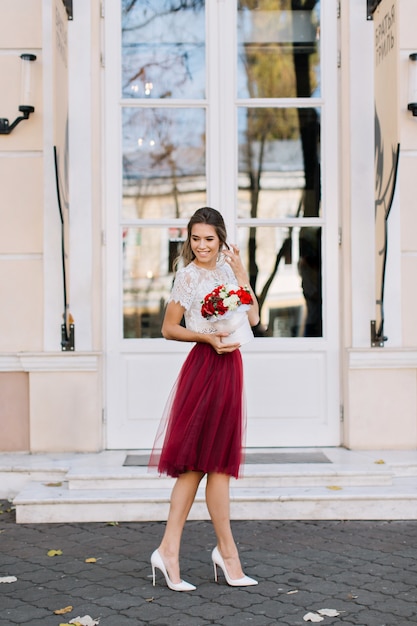 Free photo beautiful girl in marsala tulle skirt with light hairstyle walking on street. she holds  flowers and smiling to side