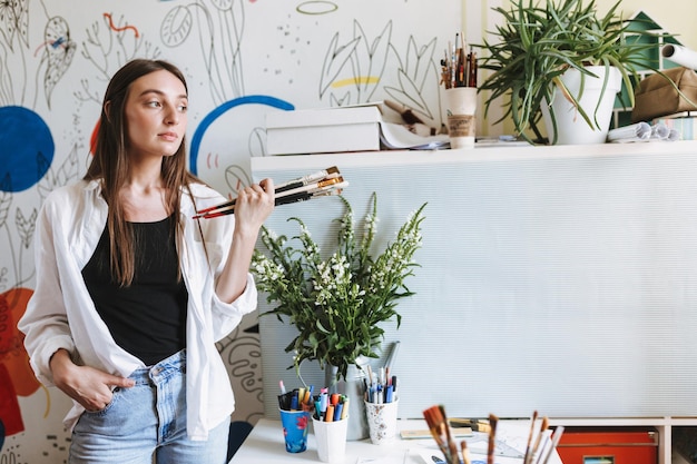 Free Photo beautiful girl near desk holding paint brushes in hand while thoughtfully looking aside with big patterns canvas on background at home