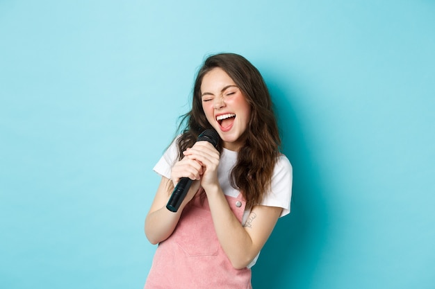 Beautiful girl singer holding microphone, singing karaoke in mic, standing over blue background.