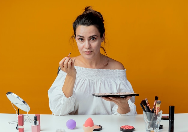 Free photo beautiful girl sits at table with makeup tools holding makeup brush and eyeshadow palette looking isolated on orange wall