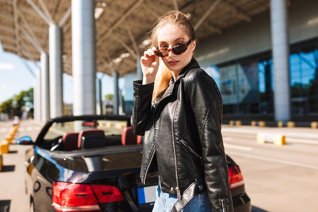 Free photo beautiful girl in sunglasses and leather jacket thoughtfully looking in camera near airport with black cabriolet car on background