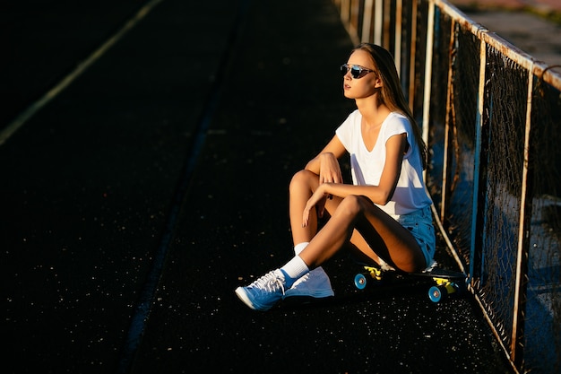 Free photo a beautiful girl in the sunglasses sitting near the fence on her skateboard on sunset.