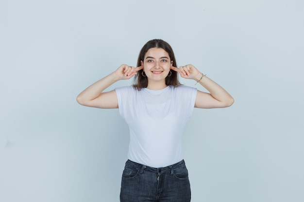 Beautiful girl in t-shirt, jeans holding fingers in ears and looking cheerful , front view.