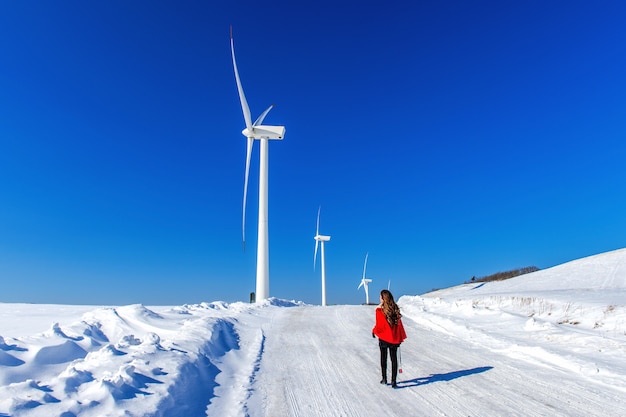 Beautiful girl walking in winter landscape of sky and winter road with snow and red dress and wind turbine