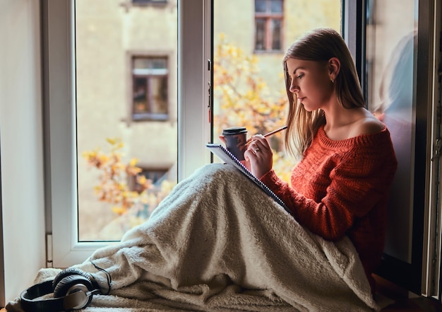 Beautiful girl in a warm sweater covered her legs with a blanket, makes notes in her notebook sitting on the window sill next to the open window