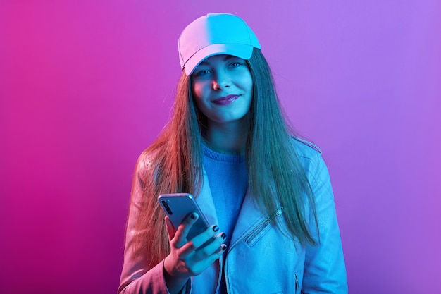 Beautiful girl wearing basket cap and leather jacket, holding modern smart phone in hands, looking smiling directly at camera