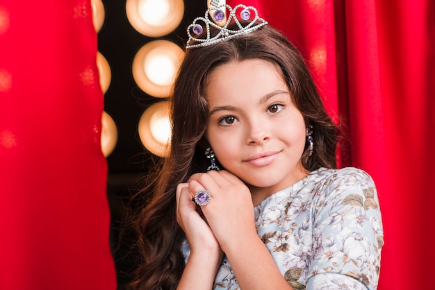 Free photo beautiful girl wearing crown standing in front of red curtain