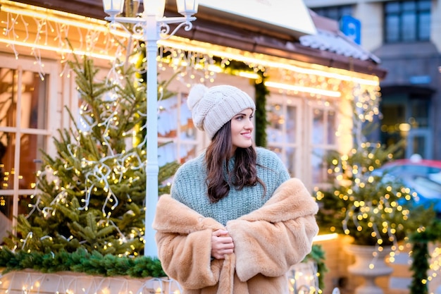 Free Photo beautiful girl wearing a winter hat and fur coat standing in evening street decorated with beautiful lights at christmas time