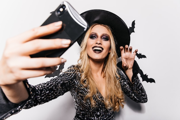 Free photo beautiful girl in witch attire making selfie at carnival. long-haired blonde lady chilling in halloween.