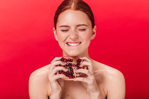 Free photo beautiful girl with fresh garnet laughing with closed eyes. studio shot of smiling amazing woman with pomegranate on red background.