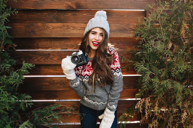 Free Photo beautiful girl with long hair in knitted hat and white gloves on wooden  surround green branches. she wears warm sweater, holds camera, looks enjoyed.