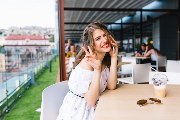 Beautiful girl with long hair sits at  table on the terrace in cafe . She wears a white dress with bare shoulders and red lipstick . She is speaking on phone and smiling to the camera.