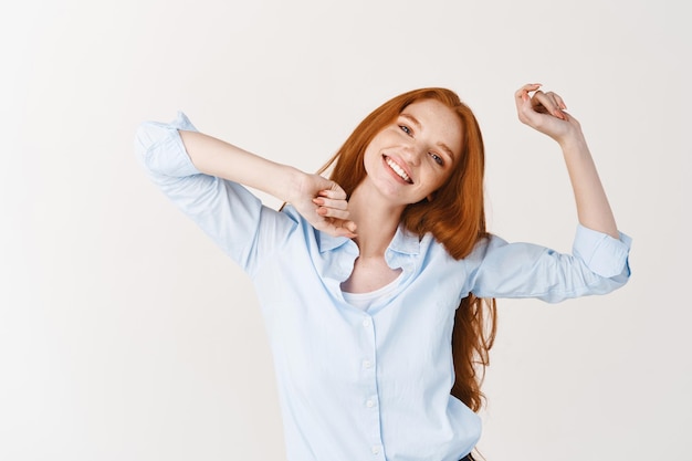 Beautiful girl with long red hair snap fingers and dancing, enjoying listening music, standing against white wall