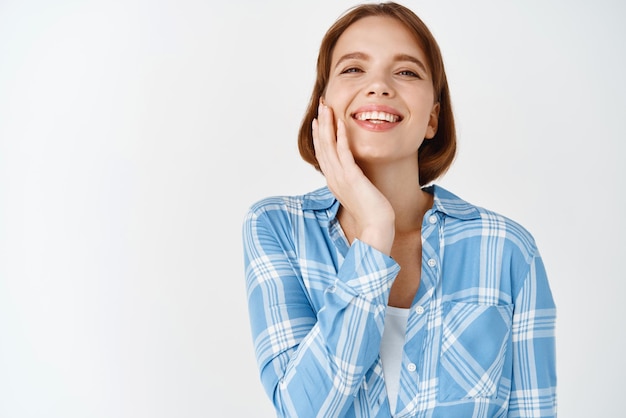 Beautiful girl with natural light makeup touching cheek and smiling happy Young woman feeling clean hydrated facial skin after applying cosmetics white background