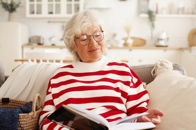 Beautiful gray haired senior European woman in stylish round eyewear enjoying reading novel, sitting on couch with book. Charming grandmother resting at home, looking through exciting textbook