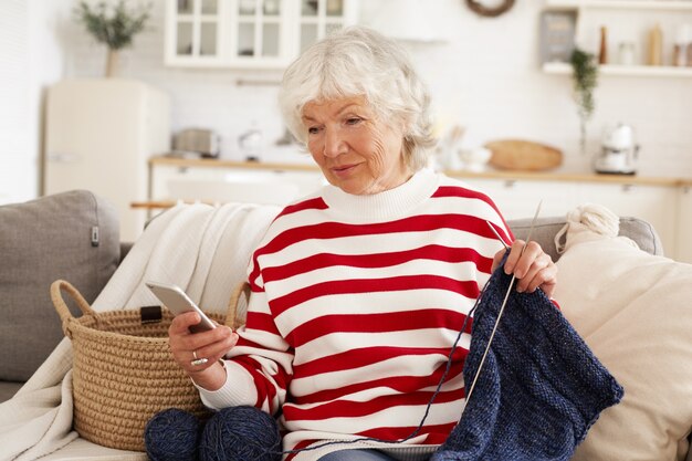 Beautiful gray haired woman on retirement spending rainy day at home sitting on couch and knitting, holding mobile, typing text message. Elegant grandmother messaging grandson online using cell phone