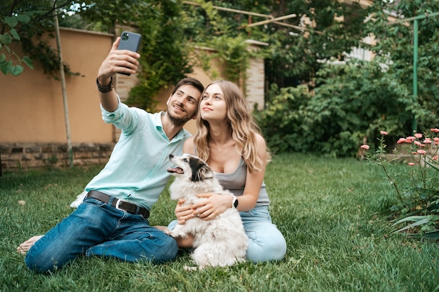 Beautiful happy couple making selfie with their lovely dog on the backyard while sitting on the grass