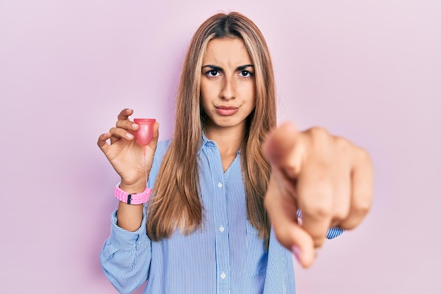 Beautiful hispanic woman holding menstrual cup pointing with finger to the camera and to you, confident gesture looking serious