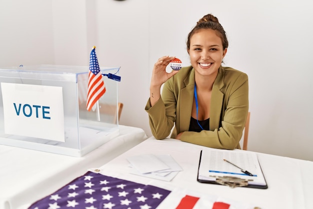 Beautiful hispanic woman at political campaign by voting ballot looking positive and happy standing and smiling with a confident smile showing teeth