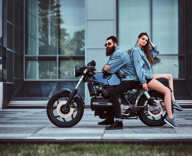 Beautiful interesting couple is sitting onthe bike in denim jacket near glass building.