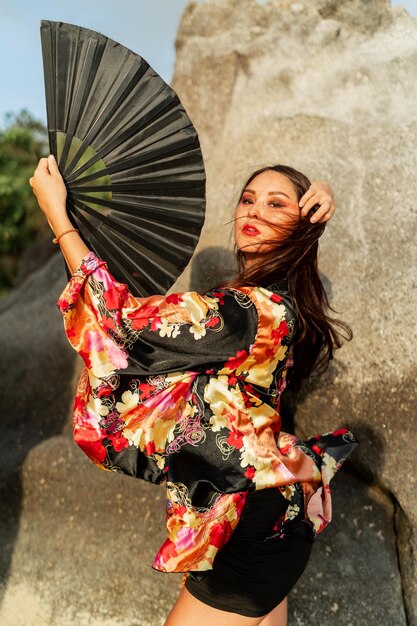 Beautiful japanese girl in stylish kimono with large fan and professional make up posing over rocks