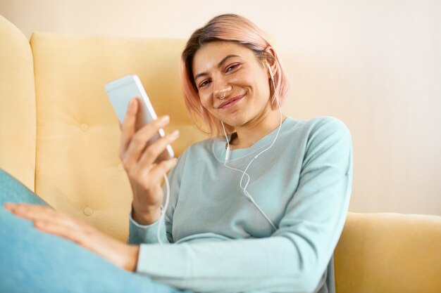 Beautiful joyful young female with pink hair and facial piercing relaxing on yellow sofa using cell phone and earbuds